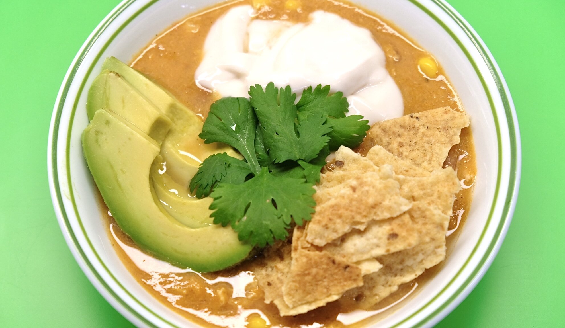 bowl of dairy free chicken tortilla soup with avocado slices and other garnishes and green background