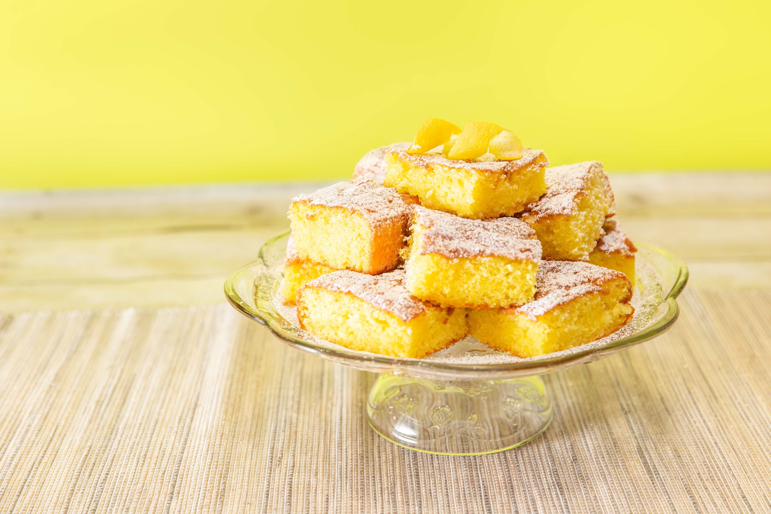 lemon cake squares on clear cake stand with yellow background