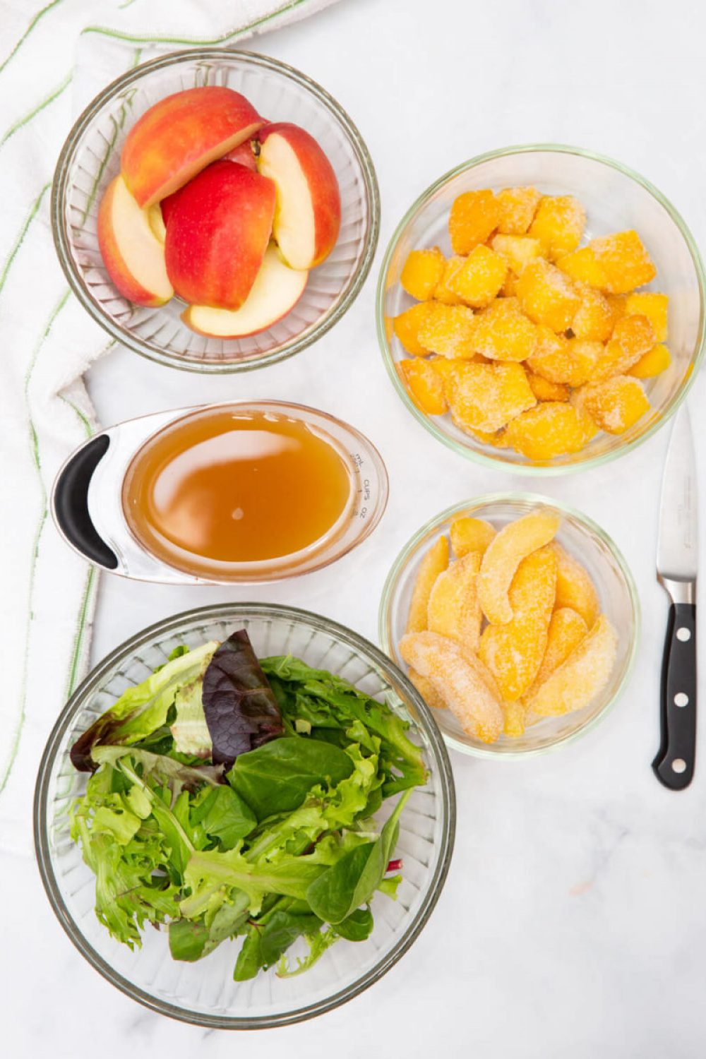 quartered apples, frozen mango, frozen peaches, apple juice and salad greens on a marble white background